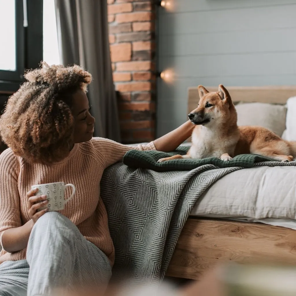 A woman sitting on the floor with a cup of coffee, smiling at a Shiba Inu resting on a bed